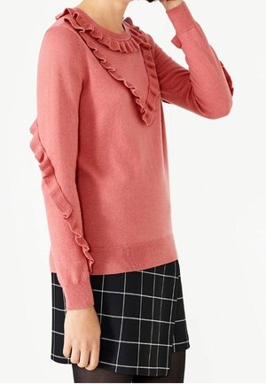 Rosa Pullover mit Rüschenausschnitt und Ärmelakzenten, aus einem weichen Material. Kombiniert mit einem schwarz-weiß karierten Rock.