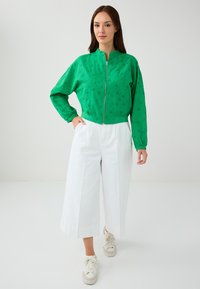 Groene ritsjack met een bloemachtig oogjesontwerp, gecombineerd met witte wijdvallende broek en beige sneakers. De stof is glad en lichtgewicht.