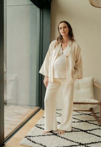 Zwangere vrouw in losse, crèmekleurige kleding staat op blote voeten op een patroonrijke kleed bij een groot raam in een moderne, minimalistische woonruimte.