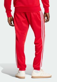 Červené atletické tepláky s tromi bielymi pruhmi pozdĺž bokov; vyrobené z hladkej látky s zužujúcim sa strihom a elastickými manžetami.