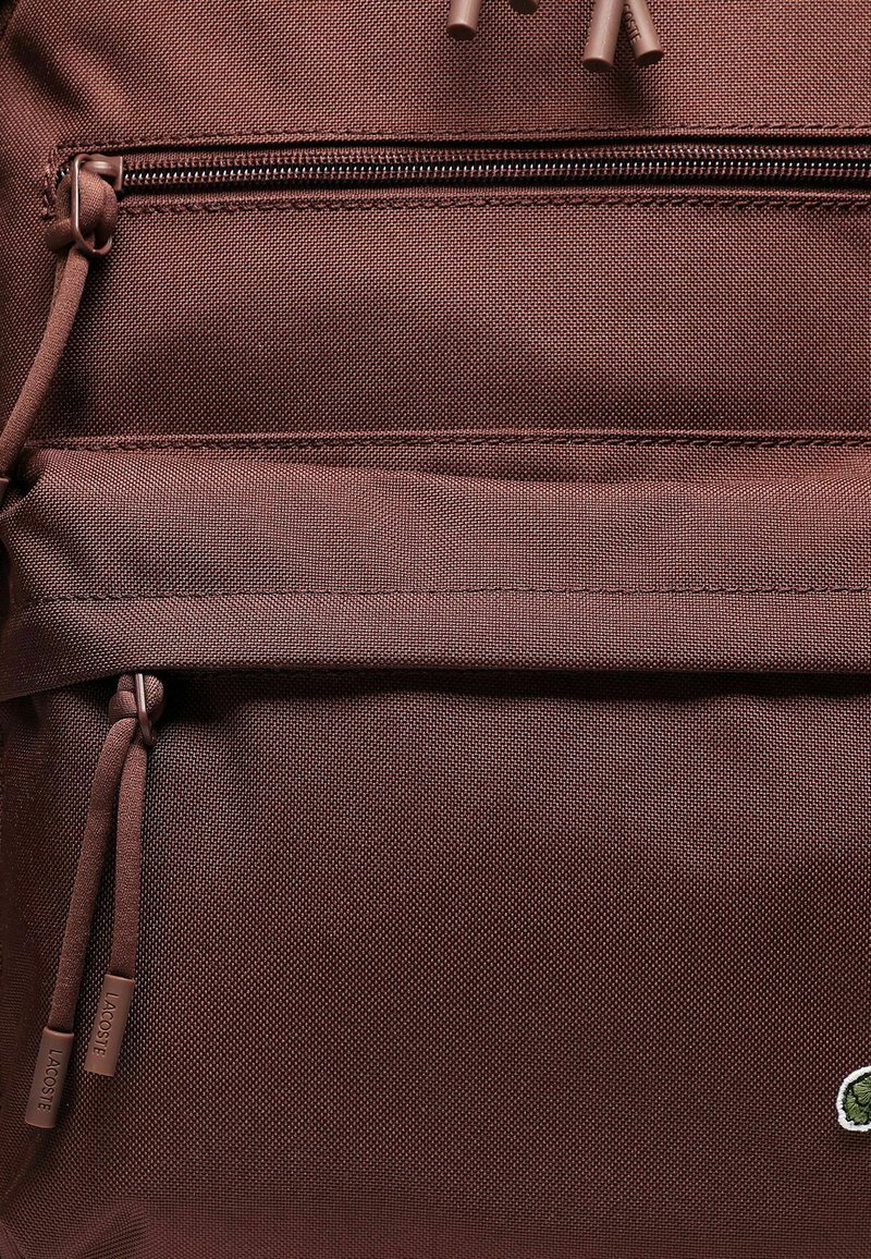 Brown fabric backpack featuring a top zipper closure, front pocket with zipper, and branded accent at the bottom. Textured exterior.