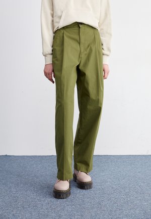 Persoon met olijfgroene broek met rechte pijpen, beige hoodie en beige schoenen met dikke zolen staand op een blauw tapijt.