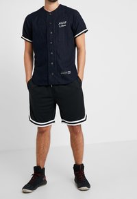 Marineblå skjorte med knapper og tynde striber, korte sorte mesh-shorts med hvide striber og sorte sportssko med røde detaljer.