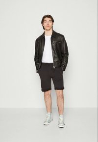 Jeune homme debout, les mains dans les poches, portant une veste en cuir noire, un t-shirt blanc, un short noir retroussé et des baskets hautes bleu clair.