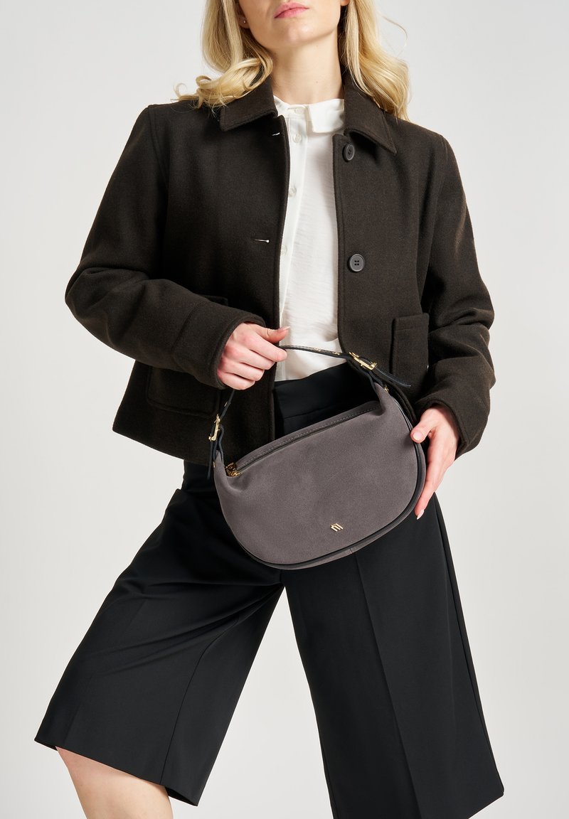 Borsa in suede grigia con forma curvata e manico in pelle nera, tenuta da una persona che indossa un cappotto scuro e una camicia bianca con pantaloni shorts neri.
