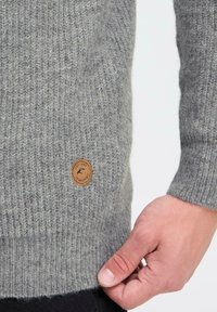 Grijze gebreide trui met ribbelstructuur, voorzien van een leren embleemlabel aan de onderkant. De mouwen en de zoom zijn ook geribbeld.