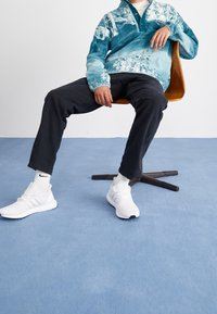 Pull polaire bleu et sarcelle avec col à demi-fermeture éclair, pantalon noir et chaussures de sport blanches, assis sur une chaise en bois sur un sol bleu.