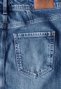 La tela de mezclilla azul claro presenta un bolsillo trasero en forma de diamante con detalles de costura. Una etiqueta de cuero marrón es visible en la cinturilla.