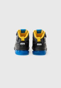 Batman-inspirerte høyde sneakers har en svart overdel, gule detaljer, blå gjennomsiktig såle, og et flaggermuslogo på hælen.