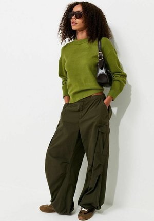 Mujer con el pelo rizado, que lleva gafas de sol oscuras, suéter verde oliva, pantalones cargo anchos, zapatillas marrones y lleva un bolso negro al hombro.