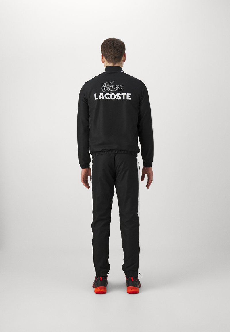 Giacca atletica nera con logo "LACOSTE" bianco e grafica del coccodrillo sulla schiena, abbinata a pantaloni neri e scarpe con accents rossi.