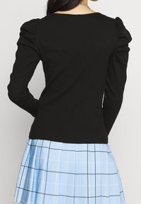 Svart långärmad topp med puffärmar, texturerad tyg och fyrkantig halsringning, i kombination med en plisserad ljusblå kjol med rutmönster.