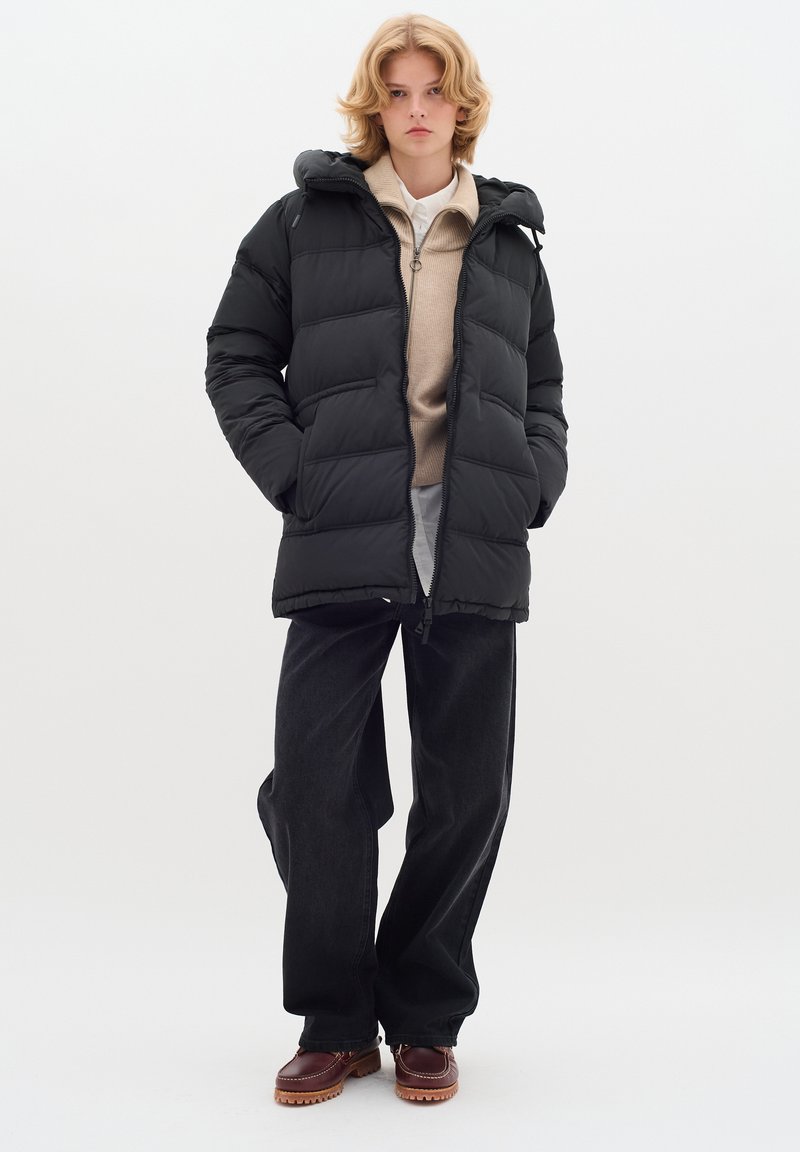 Svart puffjacka med huva och vadderad design, bärs över en beige tröja och mörka jeans. Modellen står mot en neutral bakgrund.