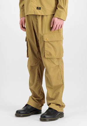 Hombre con pantalones cargo color beige con grandes bolsillos laterales y zapatos negros con cordones, de pie sobre un fondo claro y liso.