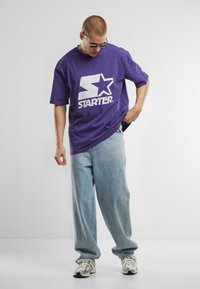 T-shirt en coton violet avec le logo "STARTER" blanc, associé à un jean ample bleu clair. Les accessoires comprennent des lunettes de soleil transparentes et des chaussures de sport.