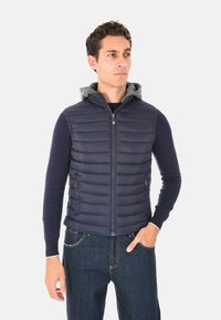 Uomo in gilet trapuntato blu navy con cappuccio grigio e maglietta a maniche lunghe scura, in piedi con una mano in tasca, che guarda di lato.