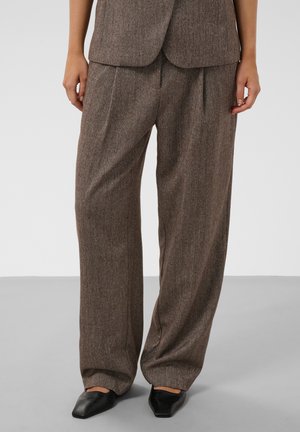 Pantalon marron à jambes larges avec motif en chevrons et plis devant, porté avec des chaussures noires à bout pointu et une veste marron assortie.