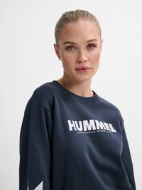 Junge Frau mit blonden Haaren in einem Pferdeschwanz, die einen dunkelblauen Pullover mit dem weißen Aufdruck "HUMMEL" auf der Brust vor einem einfarbigen Hintergrund trägt.