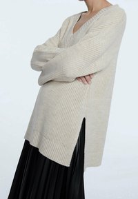 Beige gebreide trui met een diepe V-hals, losse mouwen en zijsplitten, gecombineerd met een zwarte geplooide rok. Gelaagd materiaal met geribbelde zoom.