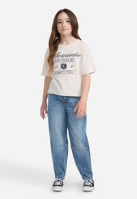 Camiseta corta beige con estampado gráfico en azul marino, combinada con vaqueros anchos de color azul claro y zapatillas en blanco y negro sobre un fondo plano.