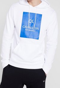 Sweat à capuche blanc avec une poche avant, arborant un graphisme rectangulaire bleu incluant le texte « CALVIN KLEIN PERFORMANCE ». Capuche avec cordon de serrage.