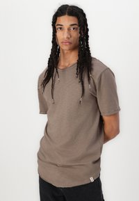 Braunes kurzärmeliges T-Shirt aus weichem Material, mit einem lässigen Schnitt, einem unvollendeten Saum und einem Etikett am unteren Ende. Subtile Struktur sichtbar.