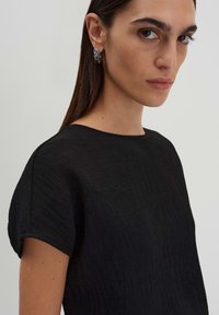 Top nero texturizzato con scollatura rotonda e maniche corte. Il modello indossa un orecchino di tono argento. Sfondo semplice, focus sui dettagli del capo.