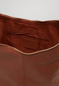 Ruskeankaltainen nahkakassi, jossa on vetoketjulla suljettava avaus, sileä ulkopinta ja kangasta vuorattu sisätasku. Kultasävyinen vetoketju.