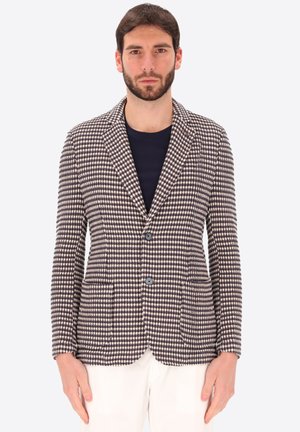 Uomo con blazer a quadri blu navy e beige sopra una camicia nera con pantaloni bianchi su sfondo semplice.