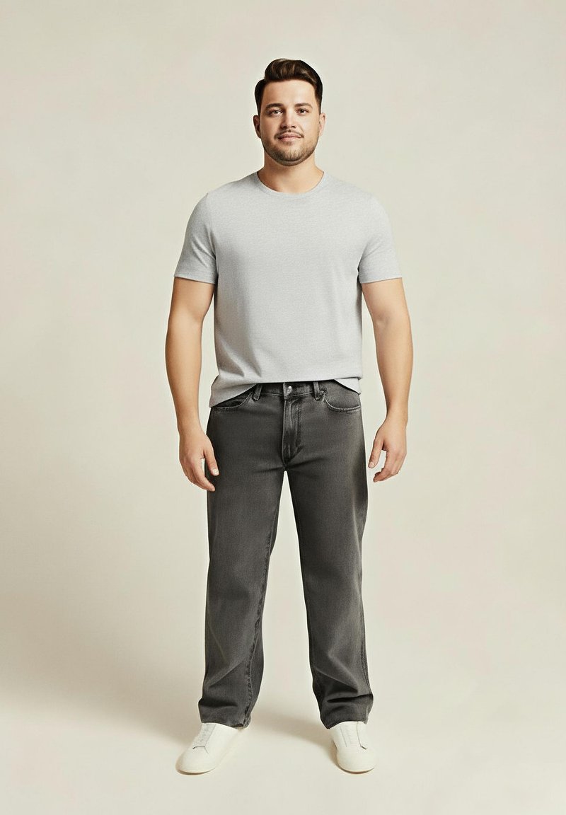Jeune homme debout, portant un t-shirt gris clair, un jean gris foncé et des baskets blanches sur un fond beige uni.