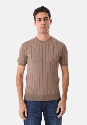 Uomo che indossa un maglione beige a costine aderente a maniche corte e jeans blu, in piedi davanti a uno sfondo bianco uniforme.