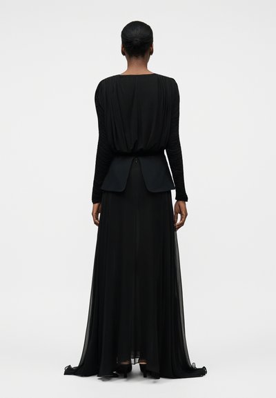Kobieta z włosami upiętymi w koczek, ubrana w długą czarną suknię z marszczonymi rękawami i wyraźną baskinką w talii, pokazana od tyłu.