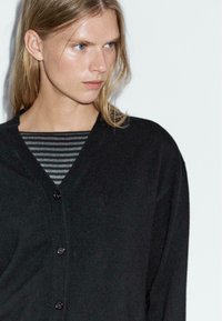 Svart cardigan med V-hals, med knappstängning och mjuk textur, lagd över en grå och svart randig skjorta.