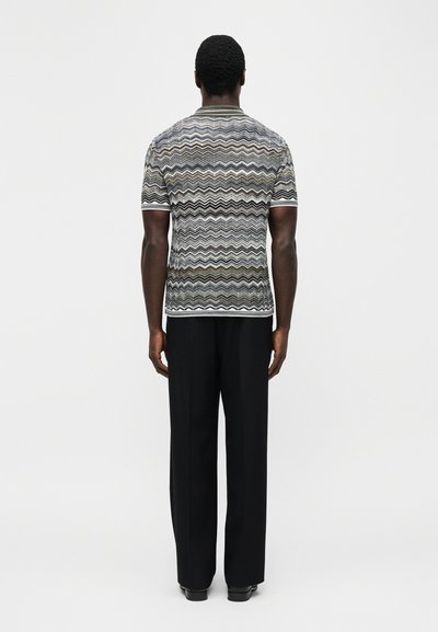 Άνδρας που στέκεται με πλάτη, φοράει κοντομάνικο πουκάμισο με σχέδιο σε σχήμα ζιγκ-ζαγκ και μαύρο παντελόνι φαρδιάς γραμμής, μπροστά σε απλό φόντο.