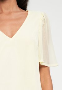 Haut jaune à col en V avec des manches courtes et transparentes. Le tissu semble léger et texturé, avec de subtiles plis sur les côtés.