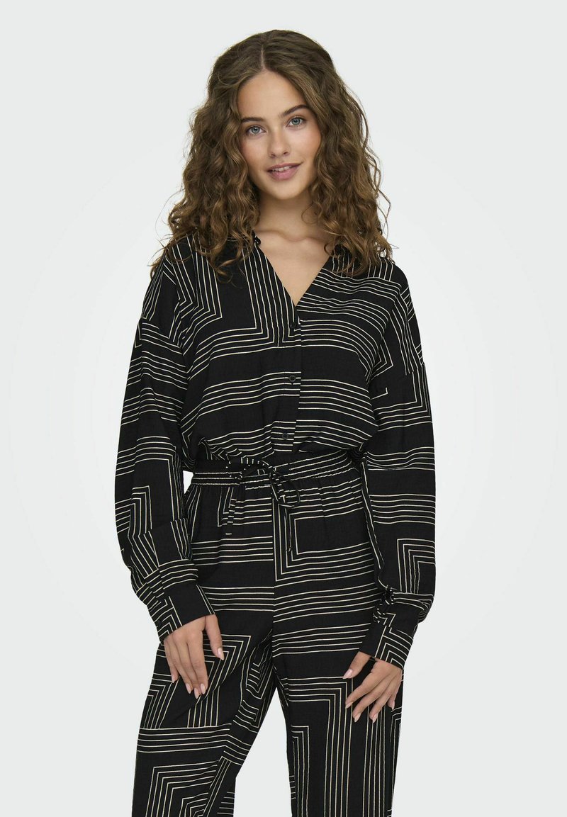 Zwarte outfit met witte geometrische lijnen, bestaande uit een blouse met kraag en elastische taillebroek. Zachte textuur, lange mouwen en een gestrikt riem.