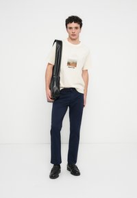 T-shirt beige avec un graphisme de paysage, pantalon bleu foncé et chaussures en cuir noir. Tenant un sac à bandoulière en cuir noir.