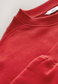 Röd sweatshirt med ribbad halsringning och muddar, mjuk textur och raka sömmar. Märkesetikett synlig nära halsringningen.