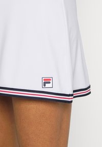 Vestido de tenis blanco con dobladillo acampanado. Presenta un ribete a rayas en azul marino y rojo en la parte inferior y un pequeño logotipo de Fila a la izquierda. Textura de tela suave.