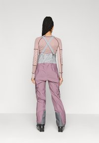 Fialové lyžiarske nohavice s sivým náprsenkou a krížovými ramienkami. Majú nákladné vrecká a zosilnené lemovanie. Materiál pôsobí odolne a vodeodolne.