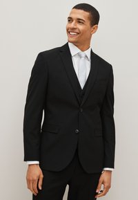 Black suit with a two-button jacket, peaked lapels, and a single chest pocket. Paired with a light tie and white dress shirt.