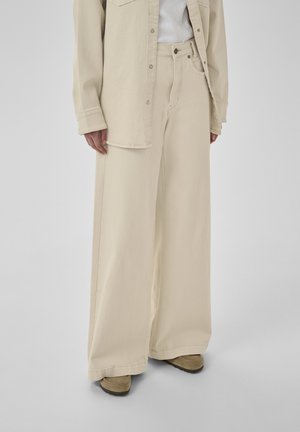 Személy bézs színű, bőszárú nadrágot, hozzáillő gombos inget, fehér felsőt és világosbarna velúr cipőt visel, egyszínű háttér előtt.