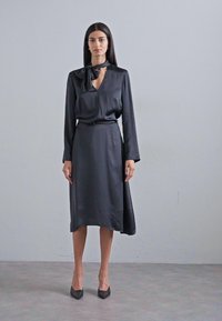 Abito in satin nero con maniche lunghe, scollatura a V e dettaglio a fiocco al colletto. Vita aderente e gonna fluttuante. Abbinato a tacchi neri.