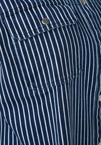 Camicia di denim con strisce verticali blu e bianche, realizzata in tessuto resistente, colletto classico, chiusura con bottoni frontale e taschino sul petto.