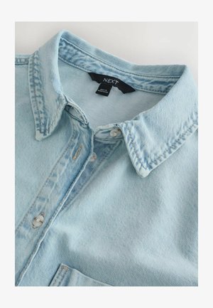 Svetlomodrá rifľová košeľa s ostrým golierom, bielymi gombíkmi a štítkom značky "NEXT" vo vnútri goliera, položená na bielej ploche.