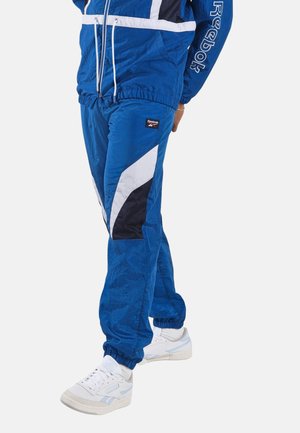 Persoon die blauwe Reebok trainingsbroek en bijpassend jack draagt met witte en zwarte accenten, gecombineerd met witte sneakers.