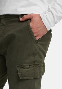 Homme portant un pantalon cargo vert olive, la main dans la poche avant, et une chemise blanche à manches longues, devant un fond uni.