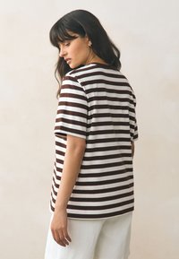 Barna-fehér csíkos rövid ujjú póló, kerek nyakkivágással és kényelmes szabással, világos színű, bő fazonú nadrággal párosítva.