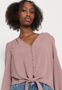 Short-sleeve, V-neck blouse in soft mauve fabric, featuring a button front and tie detail at the hem. Smooth texture with a relaxed fit.