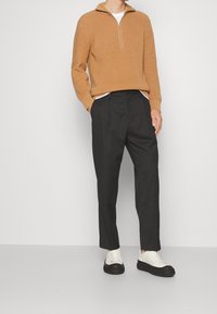 Mężczyzna w camelowym swetrze na suwak z dzianiny, czarnych plisowanych spodniach i białych trampkach z czarnymi podeszwami stoi przy gładkiej ścianie.